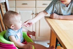 baby sitting in a high chair being fed snacks from sibling