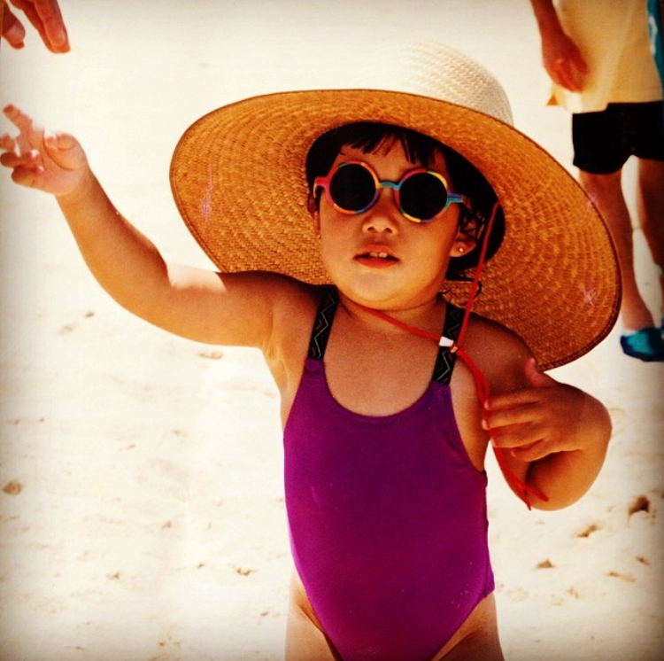 Speech Language Pathologist Amy as a toddler at the beach, with an oversized hat and round glasses