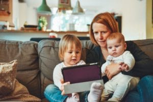 mom sitting on sofa with toddler daughter and infant daughter while looking at a tablet