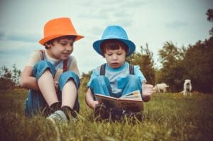 two siblings, one in a orange hat, and one in a blue hat reading a book together