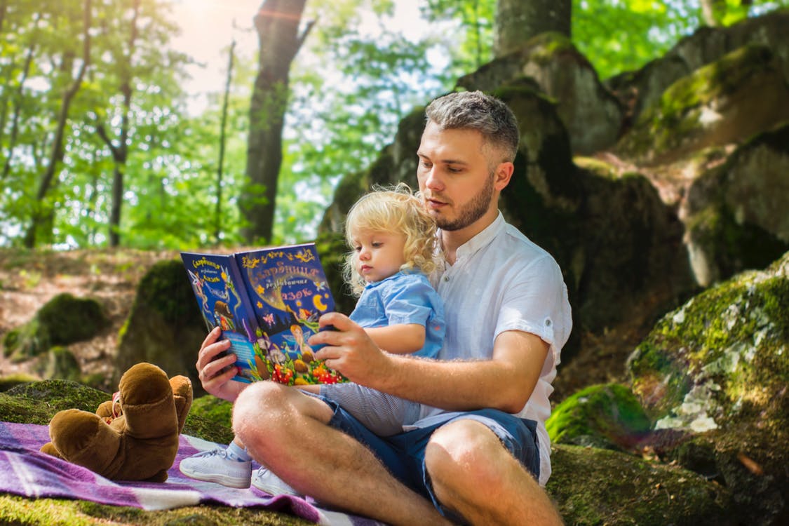father reading a book to his daughter while picnicking in the woods