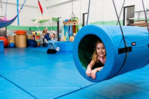 girl swinging inside a tunnel swing and another girl on the frog swing in the background in the sensory gym