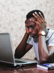 young adult male looking at his laptop frustrated with both hands on his head