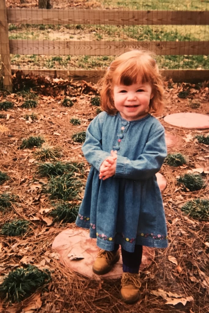 Occupational Therapist Maddy as a little girl in a denim dress standing outside with a fence in the background