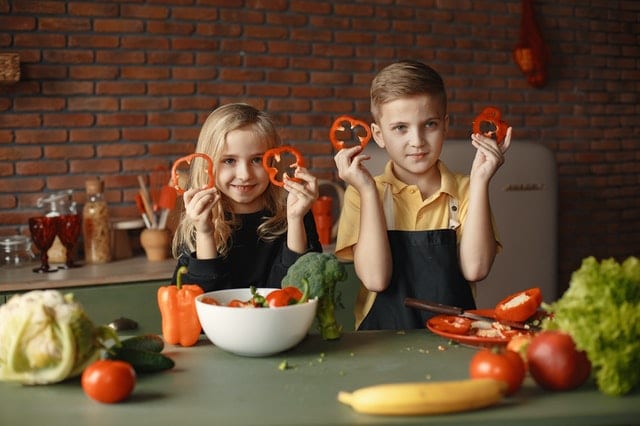 niña y niño en una cocina, sosteniendo rodajas de pimiento rojo