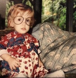 Occupational Therapist Miranda as an infant girl wearing large glasses and a red, white, and blue onesie sitting next to a blue pillow