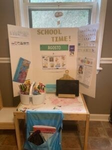 image of a children's table with a chair, ipad, and bucket of school supplie, a trifold display board with school information is set up on the table