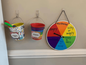 two buckets and a spinner board set up as a sensory station containing chewies, fidgets and a choice board to identify emotion