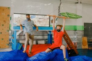 Child swings on a trapeze bar with a therapist standing behind him, leaning against a lofted platform