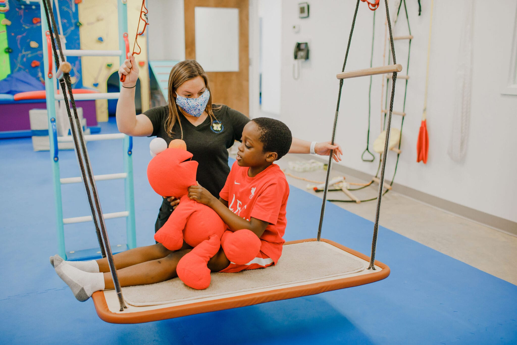 Boy sitting on the platform swing with an Elmo doll while an Occupational Therapist rocks the swing