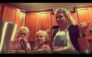 Two small blonde girls stand next to their blonde mother in a kitchen with brown cabinets