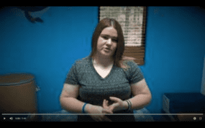white woman with shoulder length brought hair and a black striped v-neck shirt stands in front of a blue wall.