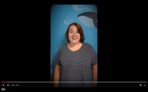 White woman with short brown hair, wearing a black and white striped shirt stands in front of a blue wall with a whale decal on it