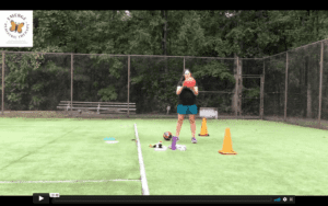 White, female therapist stand in the middle of a large green tennis court. She is holding a basketball and had cones and various equipment at her feet.