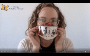 female therapist with blonde curly hairs, sits in front of a white wall, holding a colorful mask in front of her face. butterfly logo in the upper left corner of the screen