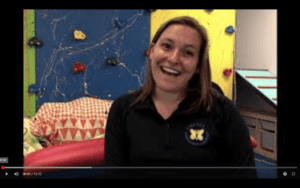 female therapist with brown hair, wearing a half zip is sitting in front of a colorful rock wall. She is smiling.