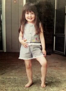 white, female child stands with one hand on hip and one knee bent. She had long brown hair, bangs, and is wearing a black and white checked top and shorts