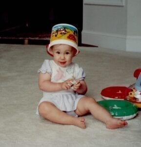 white, female, toddler in a pale summer romper sits on the floor with a colorful bucket on her head