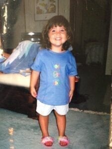white, female, toddler with short brown hair and bangs stands smiling while wearing a blue shirt and white shorts