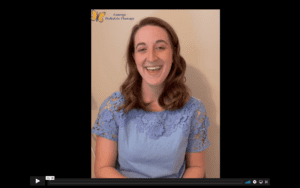 white female therapist sits in front of a beige wall. She wears a blue lacey shirt and smiles at the camera