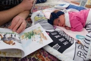 small infant with a bow lays on a black and white play mat. Her father reads a book next to her.