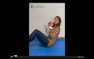 White, female therapist wearing a yellow and white striped shirt, sits in a crunch position while holding a baby doll to her chest. She looks at the camera.