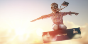 kid wearing aviator goggles flying on a suitcase with a camera around his neck